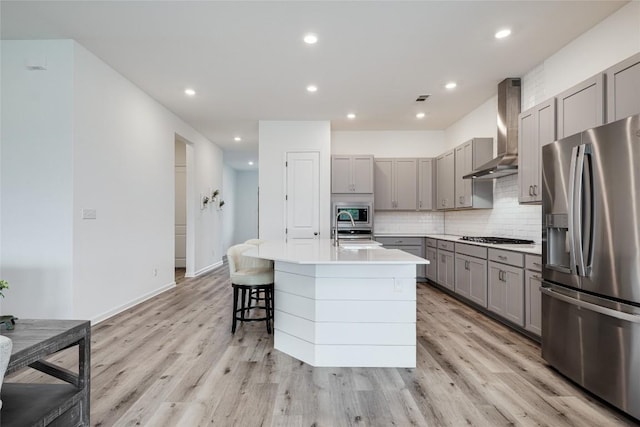 kitchen with appliances with stainless steel finishes, decorative backsplash, light hardwood / wood-style floors, a center island with sink, and wall chimney exhaust hood