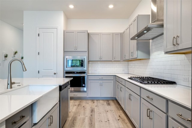 kitchen with gray cabinets, decorative backsplash, light hardwood / wood-style floors, stainless steel appliances, and wall chimney exhaust hood