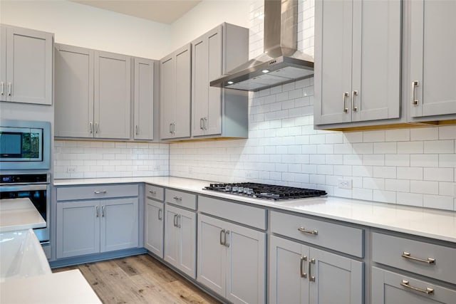 kitchen featuring light hardwood / wood-style flooring, gray cabinets, appliances with stainless steel finishes, decorative backsplash, and wall chimney exhaust hood