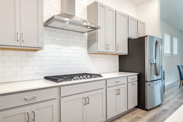 kitchen with appliances with stainless steel finishes, wall chimney exhaust hood, light hardwood / wood-style floors, and decorative backsplash