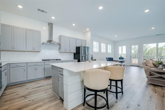 kitchen with wall chimney range hood, gray cabinets, gas stovetop, a center island with sink, and stainless steel fridge with ice dispenser