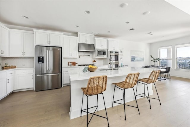 kitchen featuring a breakfast bar, a kitchen island with sink, white cabinetry, stainless steel appliances, and tasteful backsplash