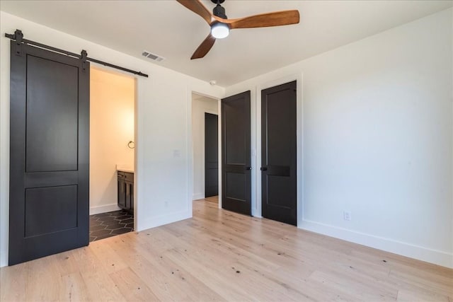 unfurnished bedroom with visible vents, light wood-type flooring, a barn door, baseboards, and ensuite bathroom