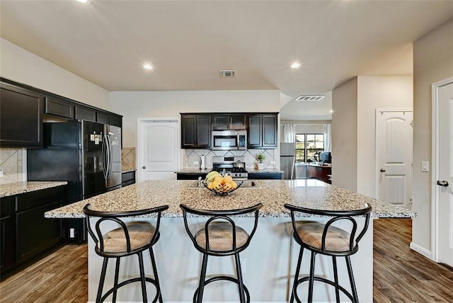 kitchen featuring a breakfast bar, stainless steel appliances, dark hardwood / wood-style floors, light stone counters, and a center island with sink