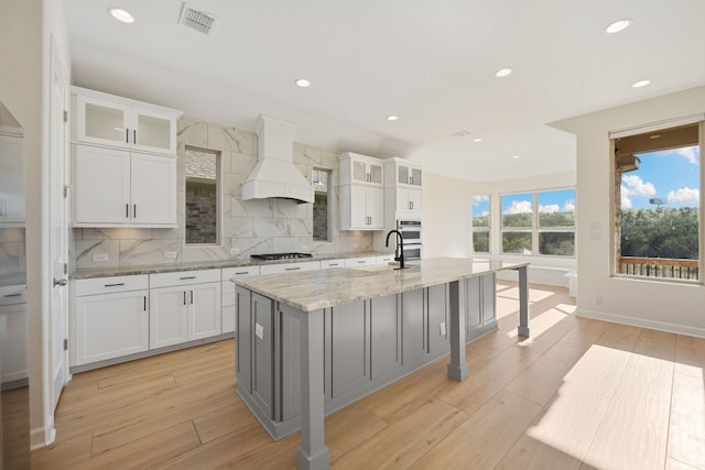 kitchen featuring a kitchen island with sink, light stone counters, custom exhaust hood, and white cabinets