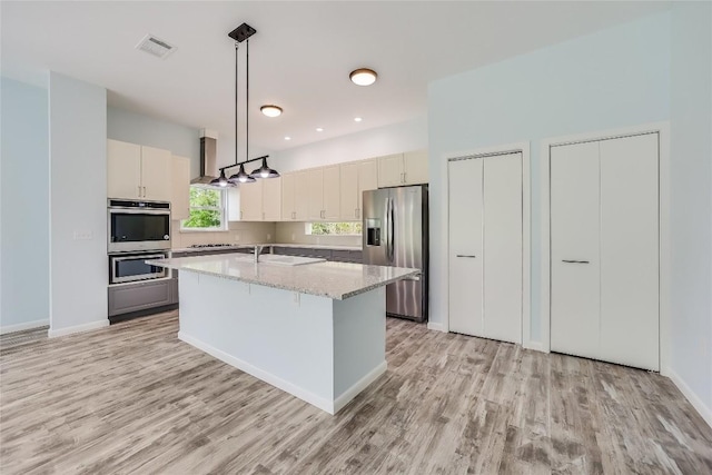 kitchen with an island with sink, hanging light fixtures, light stone counters, stainless steel appliances, and light wood-type flooring