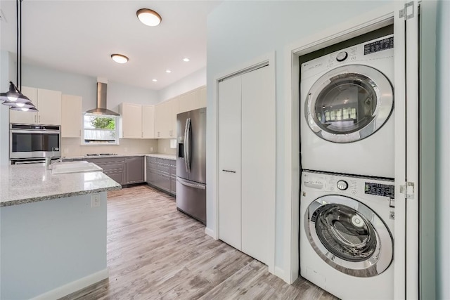 kitchen featuring wall chimney exhaust hood, stacked washer / drying machine, light stone counters, decorative light fixtures, and stainless steel appliances