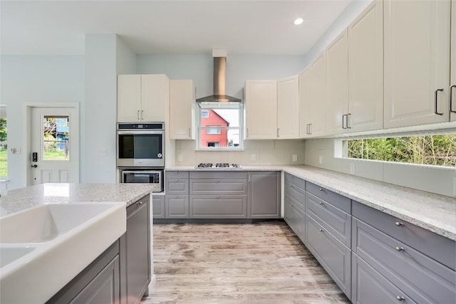 kitchen with wall chimney range hood, appliances with stainless steel finishes, gray cabinetry, light stone counters, and decorative backsplash