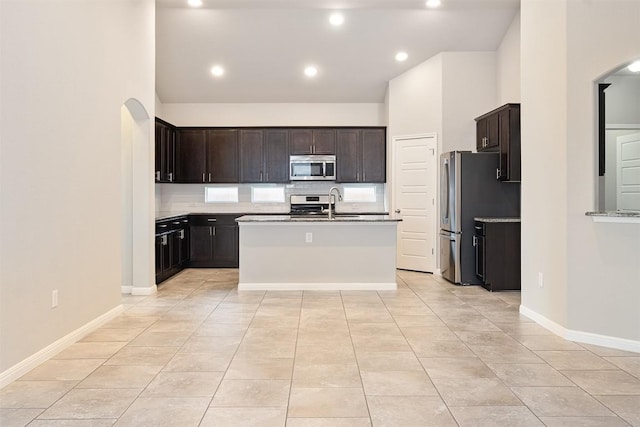 kitchen with light tile patterned floors, stainless steel appliances, tasteful backsplash, dark brown cabinetry, and an island with sink