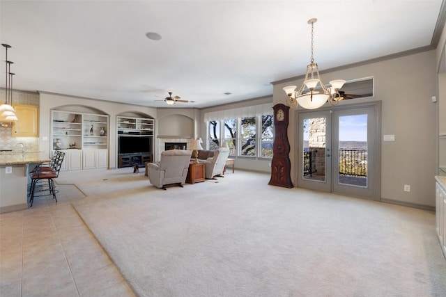 carpeted living room with built in shelves, french doors, crown molding, a fireplace, and ceiling fan with notable chandelier