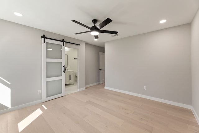 unfurnished bedroom featuring ensuite bath, a barn door, ceiling fan, and light wood-type flooring