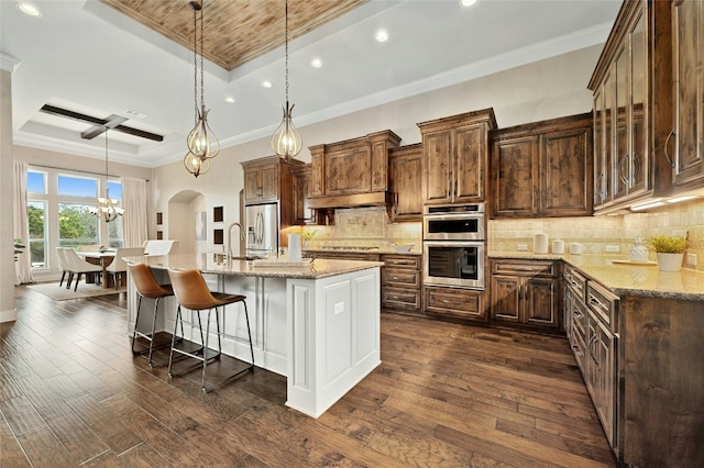 kitchen featuring appliances with stainless steel finishes, a kitchen island with sink, hanging light fixtures, a tray ceiling, and light stone countertops