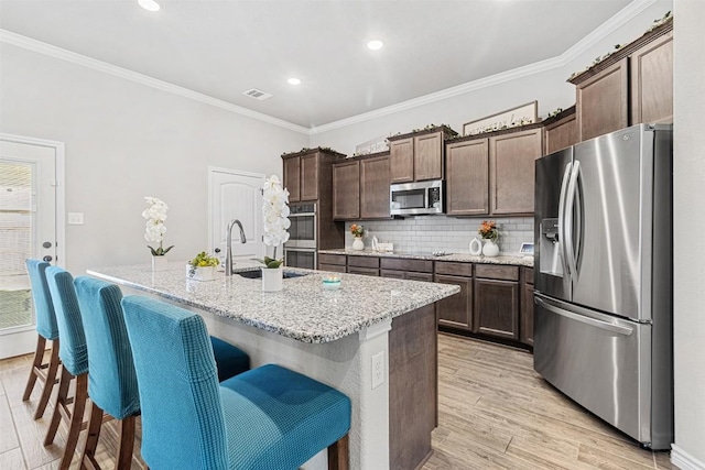 kitchen with appliances with stainless steel finishes, a kitchen island with sink, ornamental molding, light stone countertops, and light wood-type flooring