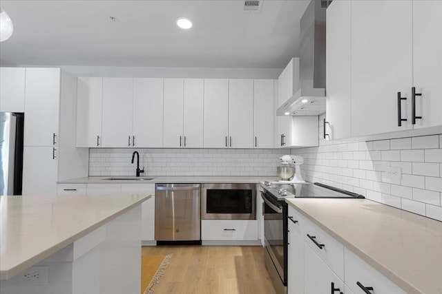 kitchen with wall chimney exhaust hood, appliances with stainless steel finishes, light wood-type flooring, white cabinetry, and a sink