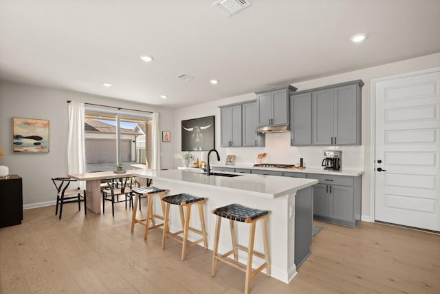 kitchen with sink, gray cabinetry, a kitchen island with sink, stainless steel gas cooktop, and a kitchen bar