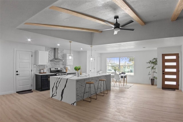 kitchen featuring wall chimney range hood, stainless steel stove, white cabinetry, hanging light fixtures, and a center island with sink