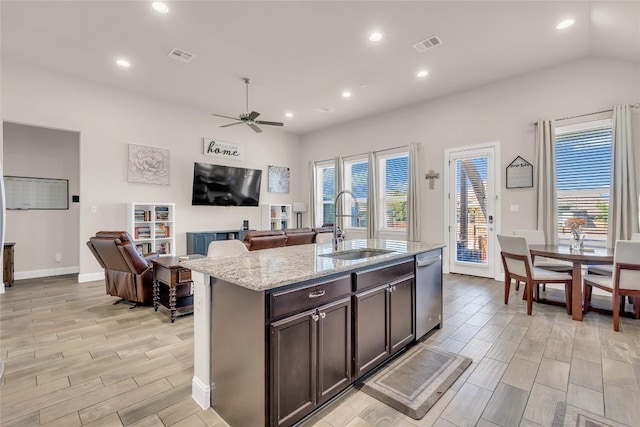 kitchen with sink, dark brown cabinets, a center island with sink, stainless steel dishwasher, and light stone countertops