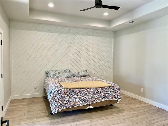 bedroom featuring wood walls, a raised ceiling, ceiling fan, and light wood-type flooring