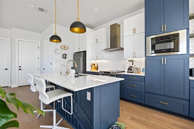 kitchen with appliances with stainless steel finishes, white cabinets, blue cabinets, decorative light fixtures, and wall chimney exhaust hood