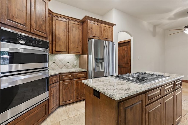 kitchen with light stone counters, a kitchen island, ceiling fan, stainless steel appliances, and decorative backsplash