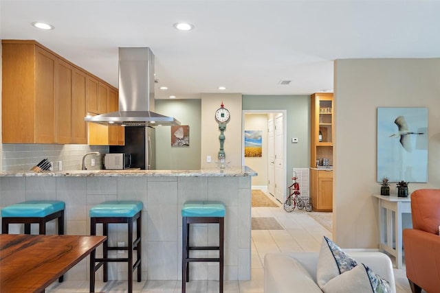 kitchen featuring a breakfast bar area, kitchen peninsula, island exhaust hood, light stone countertops, and decorative backsplash