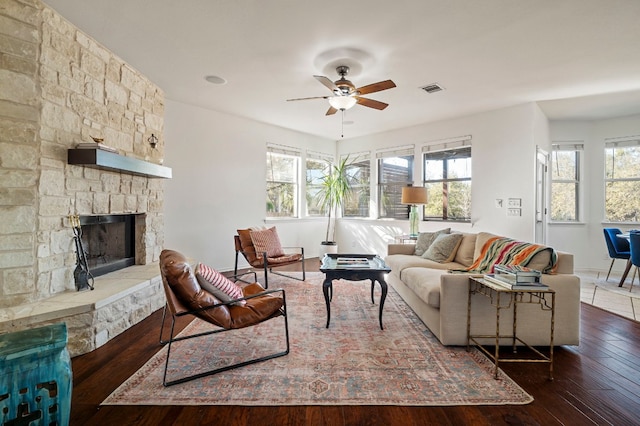 living room with a stone fireplace, dark wood-type flooring, and ceiling fan