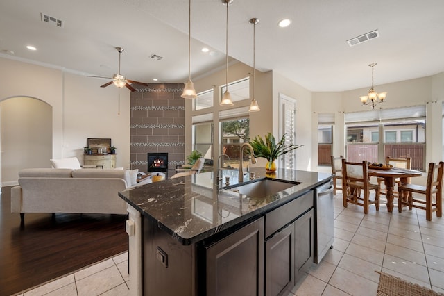 kitchen with dishwasher, sink, dark stone countertops, light tile patterned floors, and a center island with sink