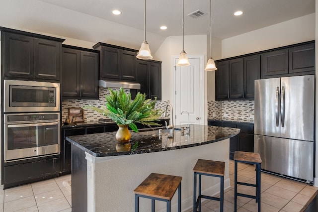 kitchen with pendant lighting, appliances with stainless steel finishes, a kitchen island with sink, light tile patterned flooring, and dark stone counters