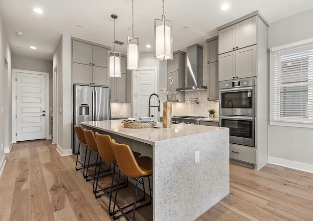 kitchen featuring wall chimney range hood, appliances with stainless steel finishes, light stone countertops, an island with sink, and light wood-type flooring