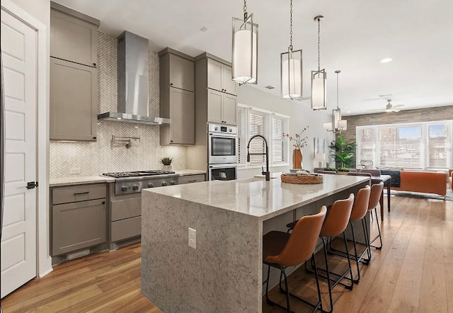 kitchen featuring wall chimney range hood, a breakfast bar, stainless steel appliances, an island with sink, and decorative light fixtures