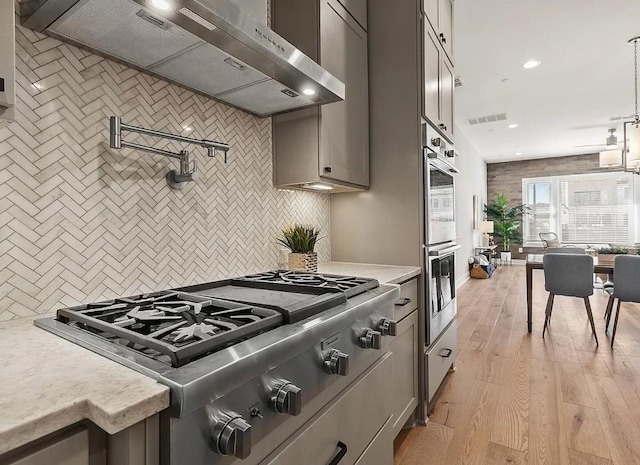 kitchen featuring gray cabinetry, light wood-type flooring, appliances with stainless steel finishes, wall chimney range hood, and backsplash