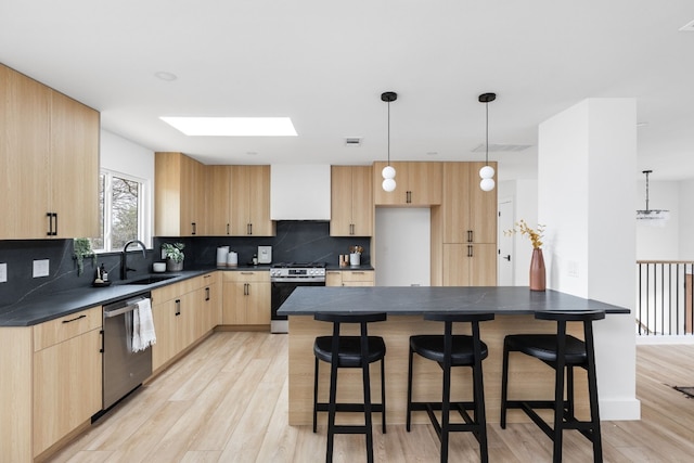 kitchen with light brown cabinetry, hanging light fixtures, appliances with stainless steel finishes, a kitchen island, and wall chimney range hood