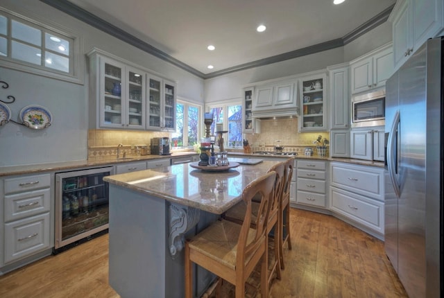 kitchen with appliances with stainless steel finishes, a center island, wine cooler, light stone counters, and ornamental molding