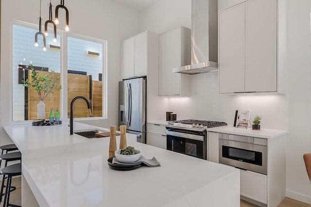 kitchen with white cabinets, a kitchen breakfast bar, hanging light fixtures, stainless steel appliances, and wall chimney range hood