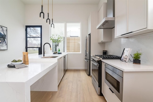 kitchen featuring a sink, wall chimney range hood, appliances with stainless steel finishes, a peninsula, and light wood finished floors