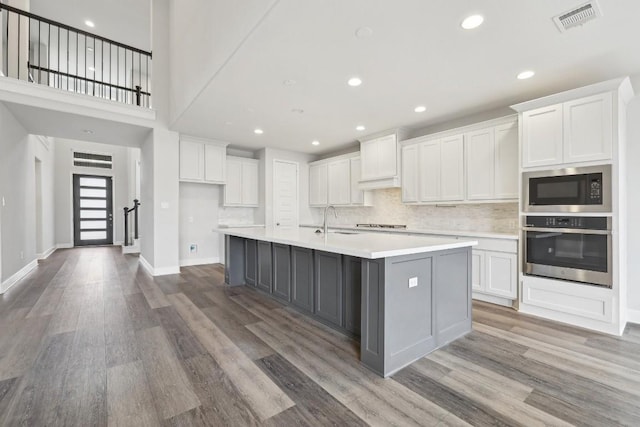 kitchen featuring a kitchen island with sink, black microwave, wood-type flooring, white cabinets, and oven