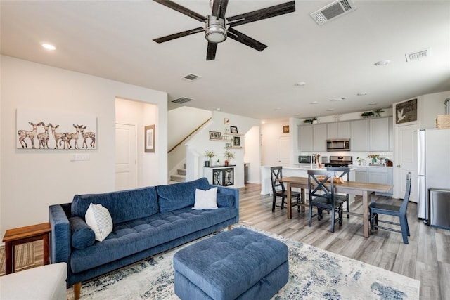 living area with visible vents, light wood-type flooring, a ceiling fan, and stairs