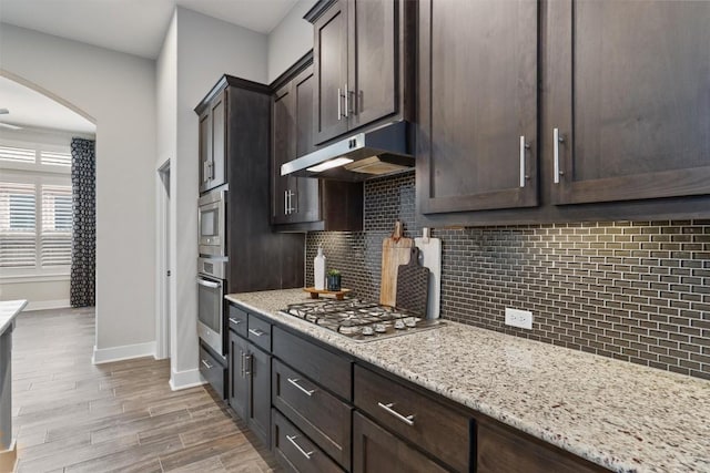 kitchen with appliances with stainless steel finishes, backsplash, light stone counters, light hardwood / wood-style floors, and dark brown cabinets