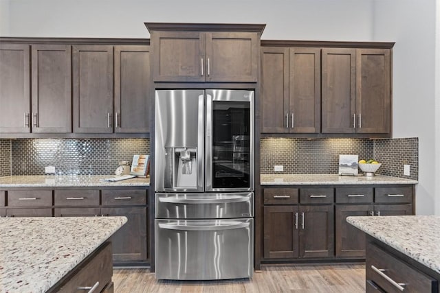 kitchen with stainless steel refrigerator, tasteful backsplash, dark brown cabinetry, light stone countertops, and light wood-type flooring