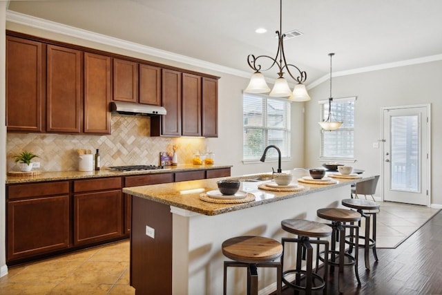 kitchen with stainless steel gas stovetop, decorative light fixtures, light stone countertops, and a center island with sink