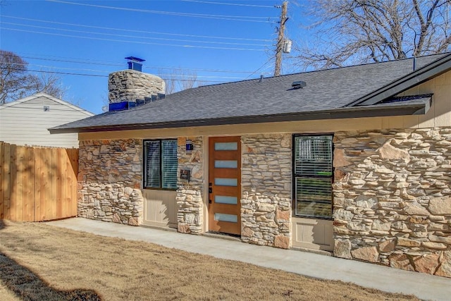 rear view of house featuring stone siding, a shingled roof, a chimney, and fence