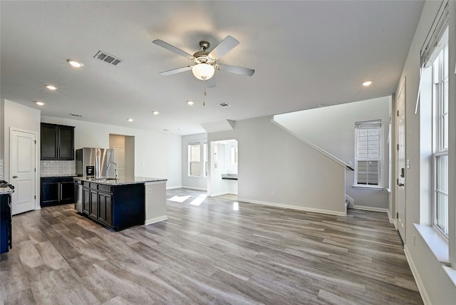 kitchen with hardwood / wood-style floors, backsplash, a kitchen island with sink, stainless steel fridge with ice dispenser, and light stone countertops