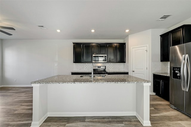 kitchen featuring sink, appliances with stainless steel finishes, wood-type flooring, light stone countertops, and an island with sink