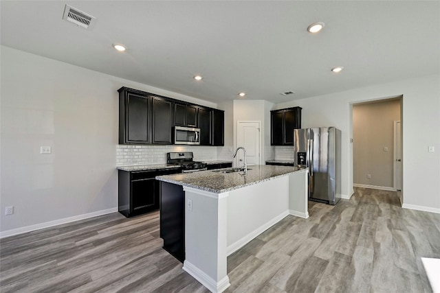 kitchen with sink, backsplash, a kitchen island with sink, stainless steel appliances, and light stone countertops