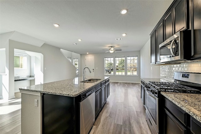 kitchen featuring sink, appliances with stainless steel finishes, an island with sink, light stone countertops, and decorative backsplash