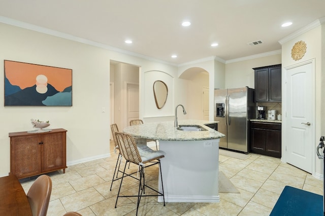 kitchen with sink, stainless steel refrigerator with ice dispenser, ornamental molding, light stone countertops, and an island with sink