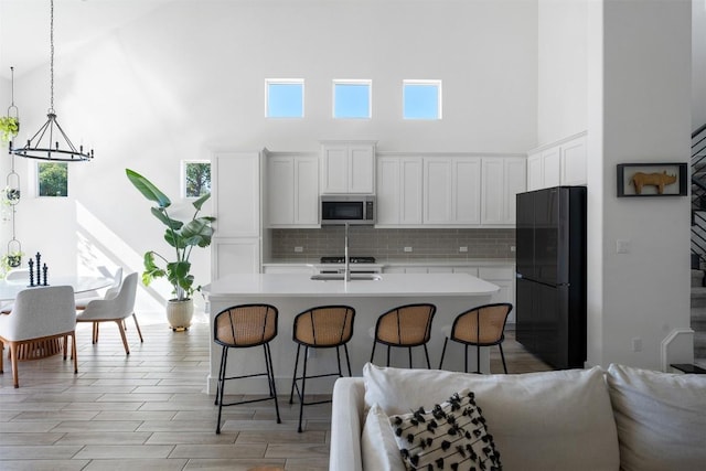 kitchen featuring backsplash, white cabinets, a center island with sink, decorative light fixtures, and black fridge