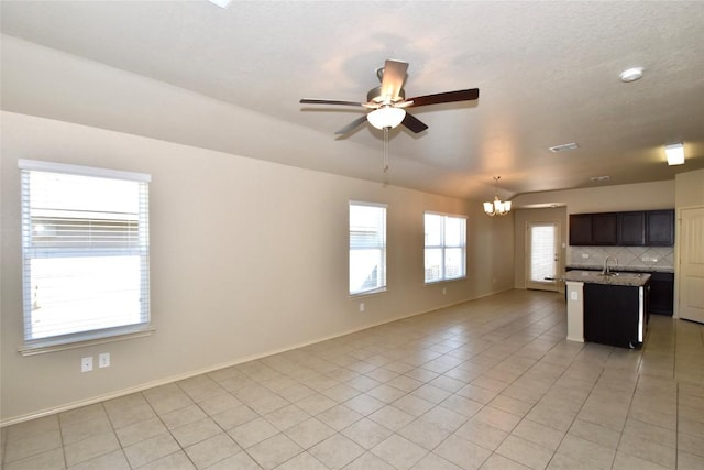 kitchen with light tile patterned flooring, tasteful backsplash, a kitchen island with sink, light stone countertops, and ceiling fan with notable chandelier