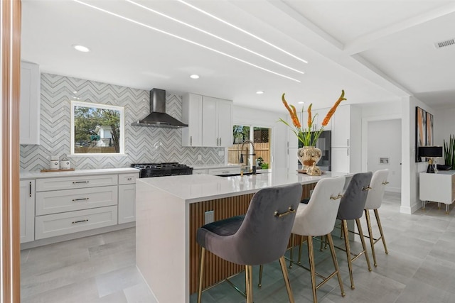 kitchen with a kitchen island with sink, a breakfast bar area, white cabinets, and wall chimney exhaust hood