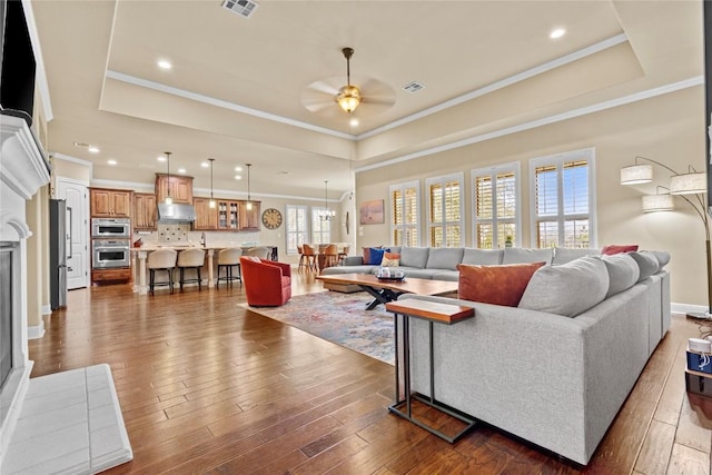 living room with dark wood-type flooring, ornamental molding, a tray ceiling, and ceiling fan with notable chandelier